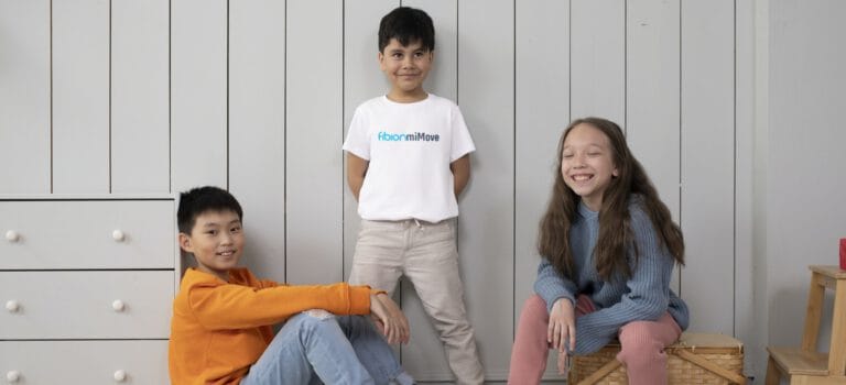 Three children are posing indoors against a white paneled wall. One child stands in the middle wearing a white shirt, and two children are seated on either side. A chest of drawers is on the left.