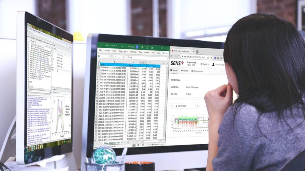 A woman is looking at two computer monitors while monitoring sleep and physical activity with an accelerometer.