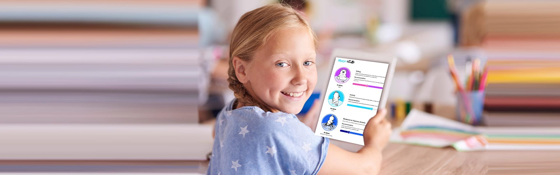 A smiling young girl showing a Fibion Kids paper with colorful icons in a classroom setting.