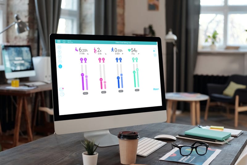 A computer screen displays a colorful graphic of stick figures and Fibion reports, placed on a desk with a coffee cup, notebook, and glasses.