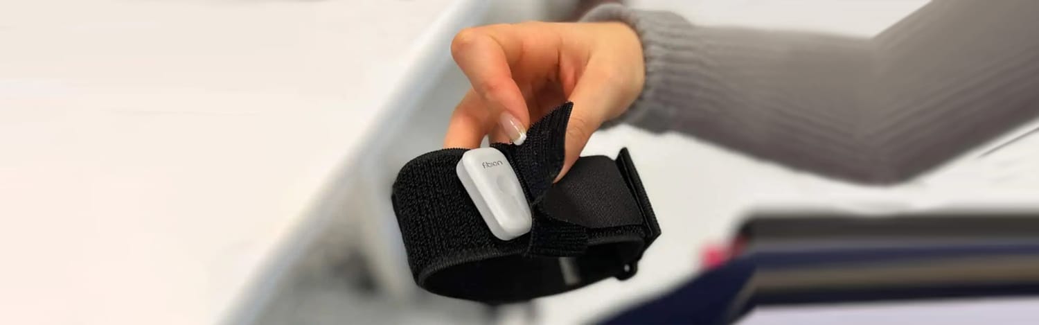 A person holds a black wrist strap with a small white electronic gadget attached to it. The person's hand is partially in the frame, and the background is blurred.