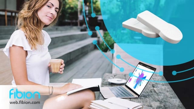 A woman sits outdoors with a laptop and coffee, surrounded by fitness equipment. The Fibion logo is tucked in the corner, hinting at her connection to Fibion SENS Cloud for analyzing physical activity data.