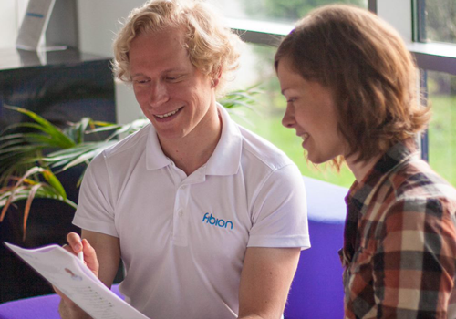 Two people are sitting together, examining a document and smiling. One wears a white shirt with a logo, the other a plaid shirt. In the background, next to the plant, they discuss menu elements that enhance their project's appeal.