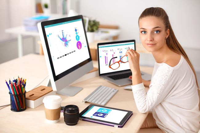 A woman sits at a desk with a desktop computer, laptop, and tablet displaying charts and graphics. The workspace includes a pen holder, coffee cup, and camera lens—alongside her Fibion Gen2 device for precise activity tracking.