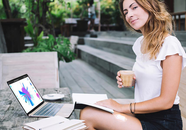 A woman sits outdoors at a wooden table with a laptop and notebook, her coffee cup in hand. She contemplates new SEO keywords to enhance her website's visibility.