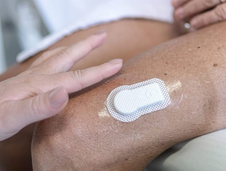 A close-up of a person's knee with a medical adhesive patch applied, highlighted by someone pointing to the patch. MM1