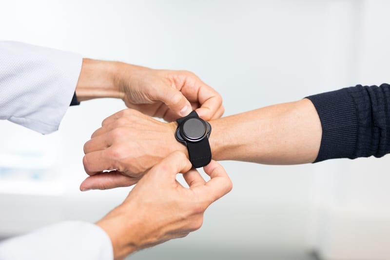 A person in a lab coat meticulously adjusts a black smartwatch on another person's wrist, ensuring its HRV tracking is optimized.