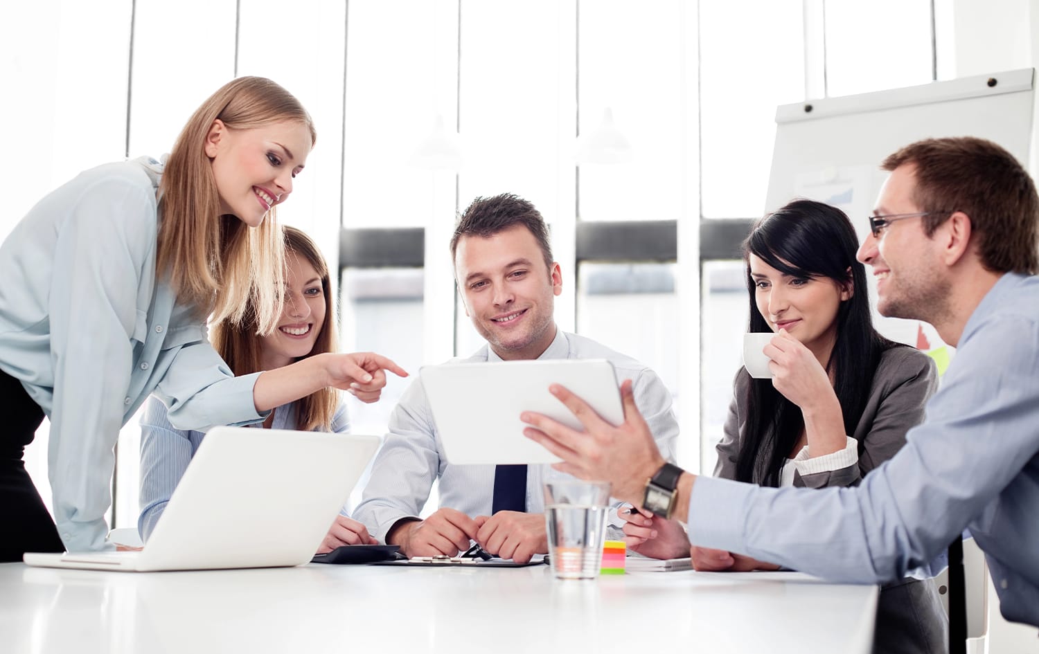 A group of five people in business attire, gathered around a table with laptops, a tablet, and Fibion software flashed on the screen, engaged in discussion in a bright office setting.