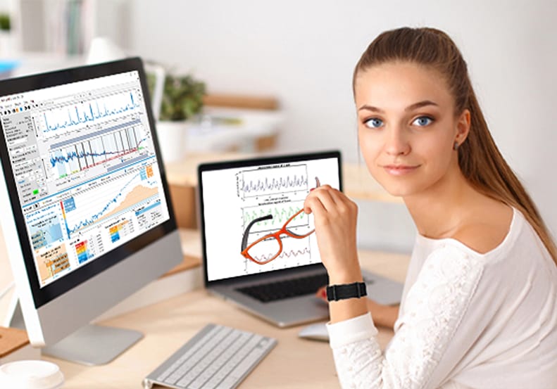 A person sits at a desk surrounded by computer screens showcasing charts and graphs, holding a pair of glasses. They gaze confidently at the camera, embodying the analytical expertise of Fibion Solutions in circadian rhythm research.