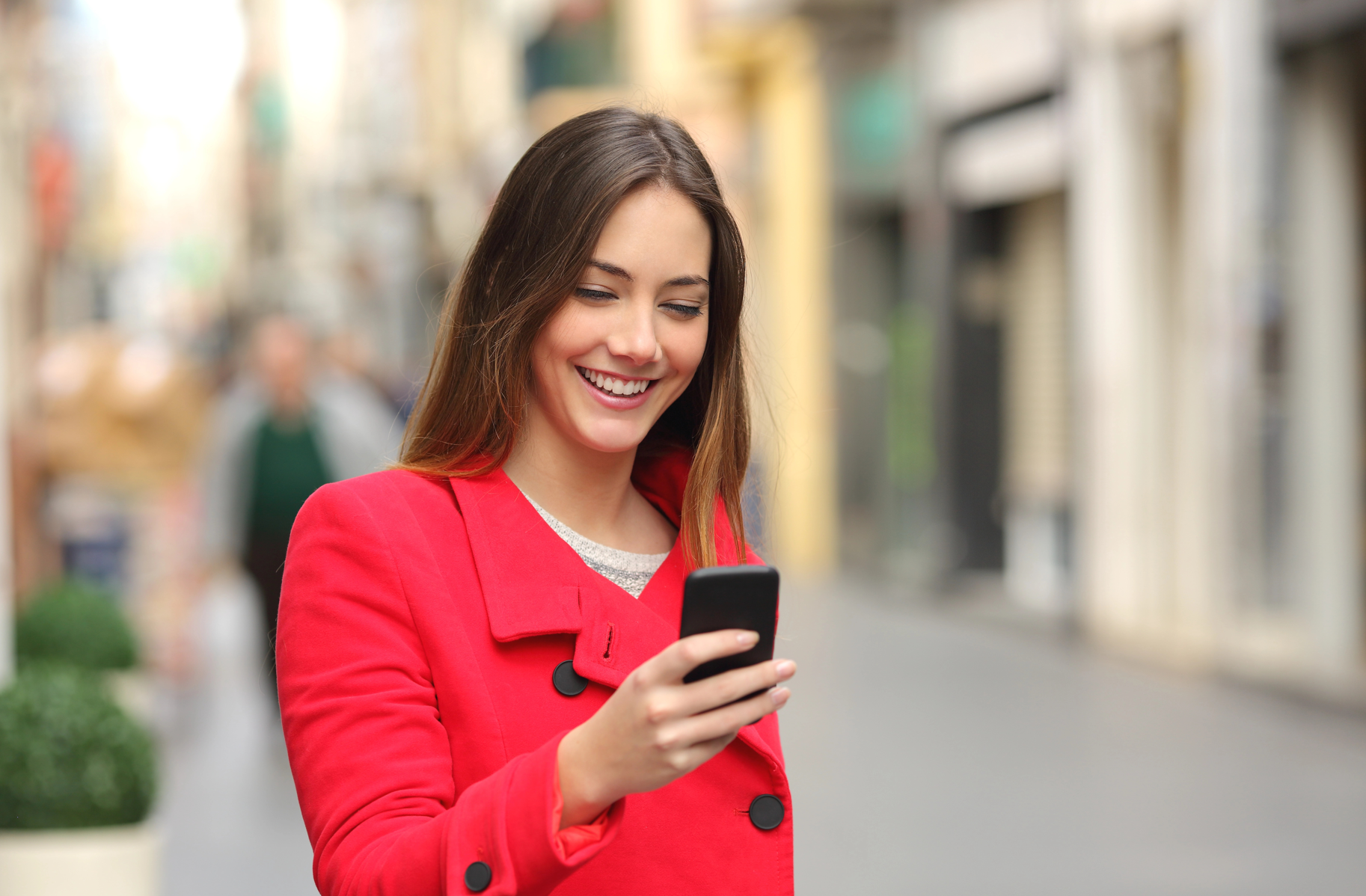 A woman in a red coat is smiling at her smartphone, tracking her steps with SENS Motion, while standing on a city street.