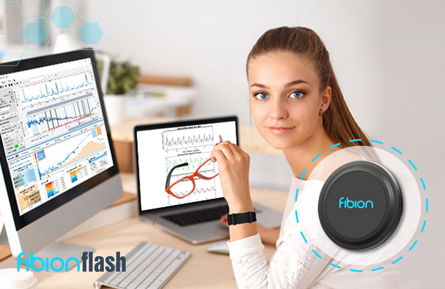 A person sits at a desk with two screens displaying graphs and HRV data. They hold glasses in one hand, while a Fibion Flash device is prominently shown.