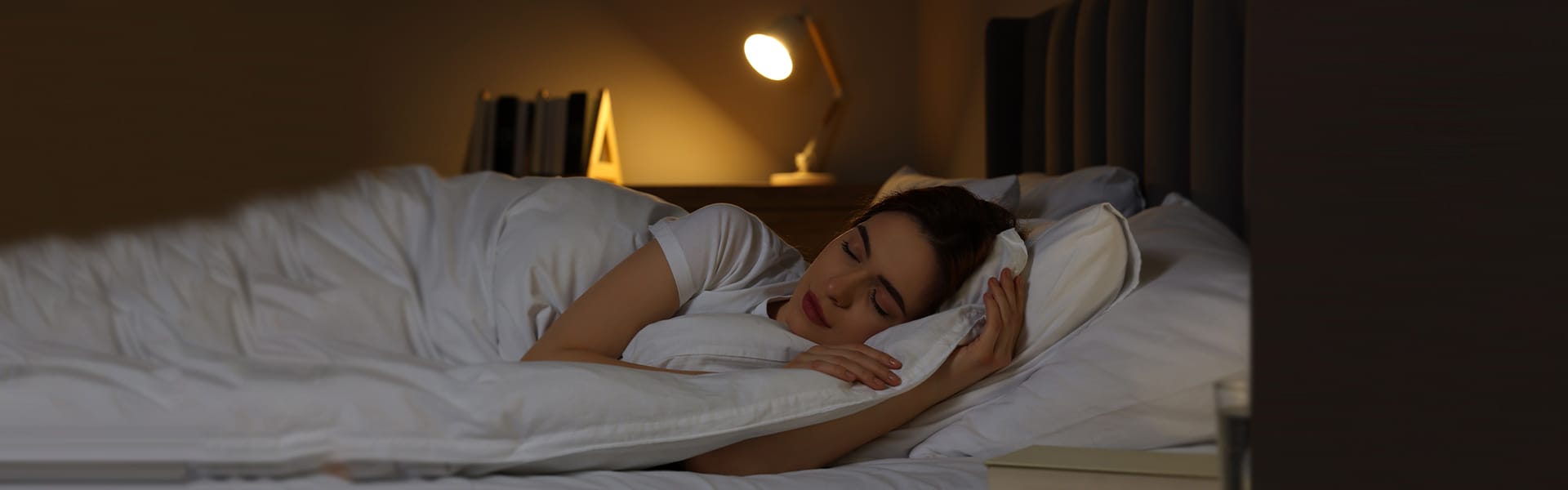 A person sleeps peacefully in bed under a white duvet, with a Fibion Krono lamp casting a gentle glow on the bedside.