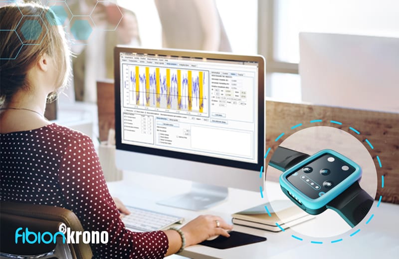 A person sits at a desk using a computer with graphic data displayed from the Fibion Krono. An inset shows a close-up of the wearable fitness tracker, seamlessly integrating health insights. The text reads "fibion krono.