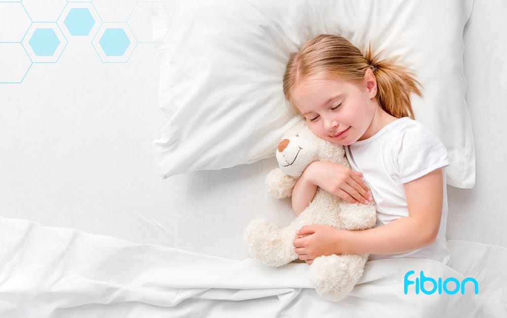 A young girl peacefully sleeps in a bed, hugging a white teddy bear on crisp white sheets. The backdrop is adorned with light blue hexagons and features a "fibion" logo in the bottom right. This serene scene subtly highlights the importance of recovery monitoring for pediatric patients.