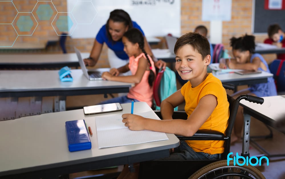 A boy in a wheelchair is engaged in writing at a desk in a classroom designed to accommodate diverse needs, while the teacher assists other students, fostering an inclusive environment for children with long-term health conditions.