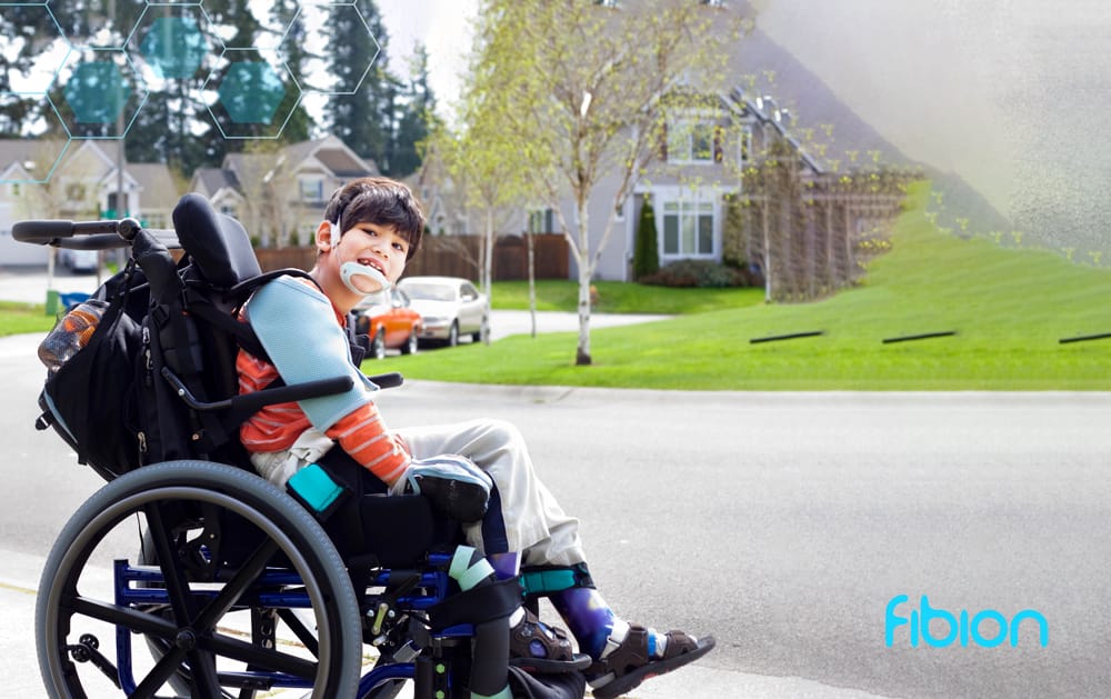 A young boy in a wheelchair, embodying the spirit of physical activity, holds a device in his mouth on a suburban street. He wears braces and carries a backpack, symbolizing how children with mobility limitations can actively engage with their surroundings. Houses and parked cars are visible behind him.